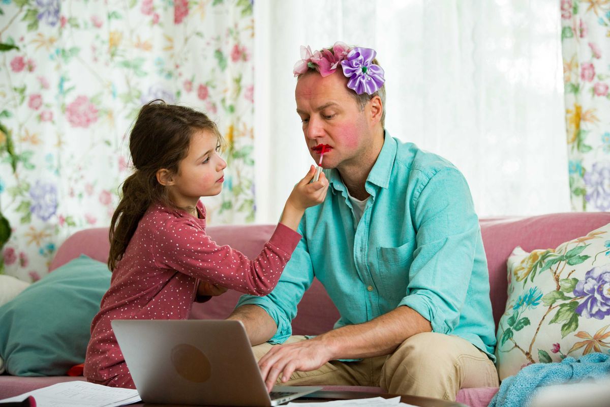 Child playing and disturbing father working remotely from home. Little girl applying makeup. Man sitting on couch with laptop.