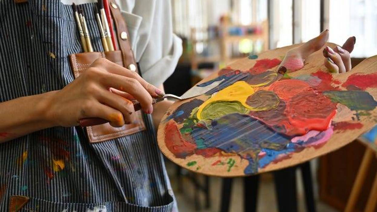 Close-up image of a young creative artist's hands smeared with watercolors holding a spatula mixing color on a palette.