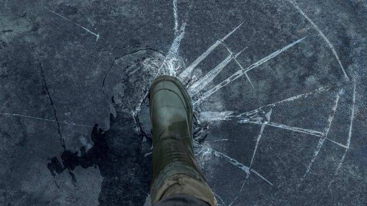 Close up of a man's winter boot pressing on the frozen surface of a lake. The ice is cracking and fracturing under the weight of his step.
