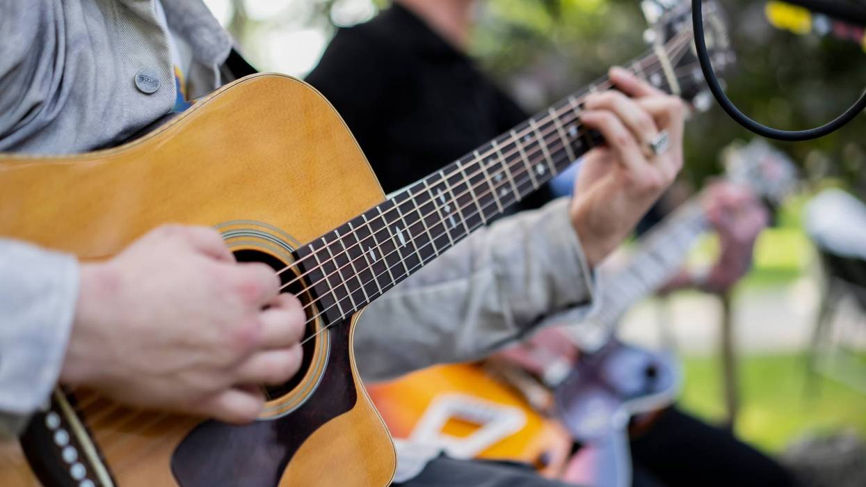 Close-up of a musician playing an acoustic guitar, focusing on the hands and strings, with other musicians blurred in the background during an outdoor performance.