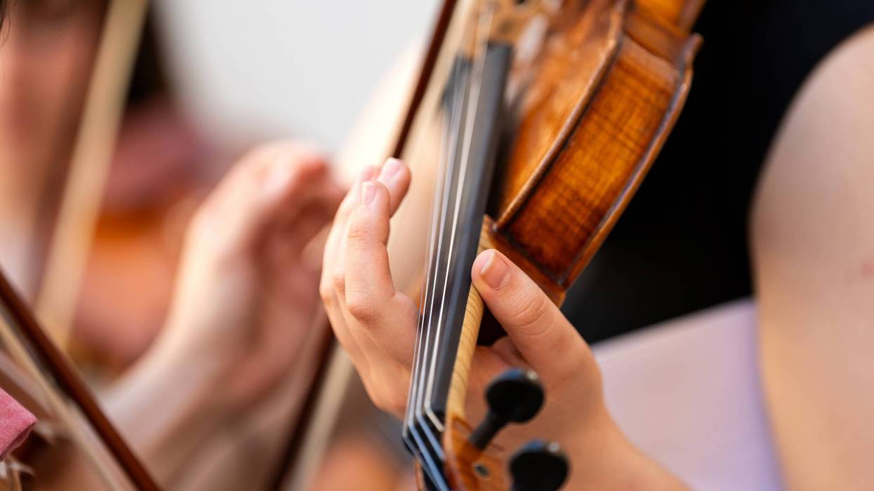 Close-up of hands playing a wooden violin, fingers pressing strings; another musician blurred nearby.