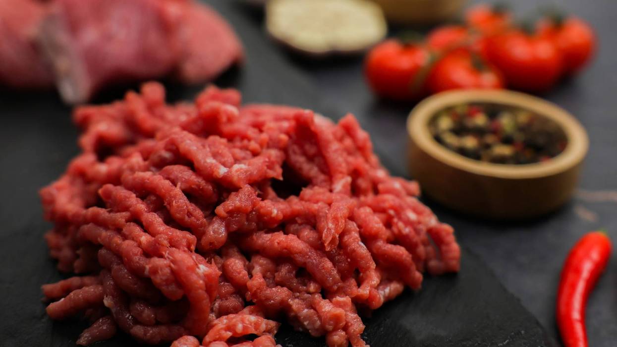 Close-up of raw red ground beef minced in a meat grinder lying on a black board, surrounded by out-of-focus cut meat, vegetables and seasonings in wooden bowls. The concept of diet, proper nutrition, natural products.
