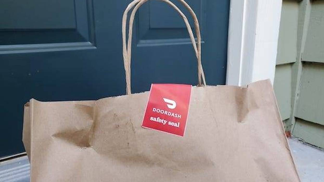 Close-up of the top of a brown paper bag, with handles and a red and white DoorDash safety seal sticker, photographed near a front door