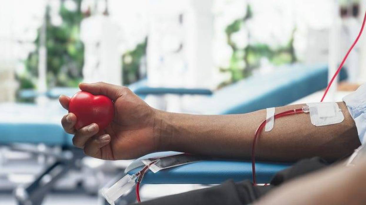 Close Up Shot Of Hand Of Male Blood Donor With an Attached Catheter. Black Man Holding Heart-Shaped Red Ball To Pump Blood Through The Tubing Into Bag.