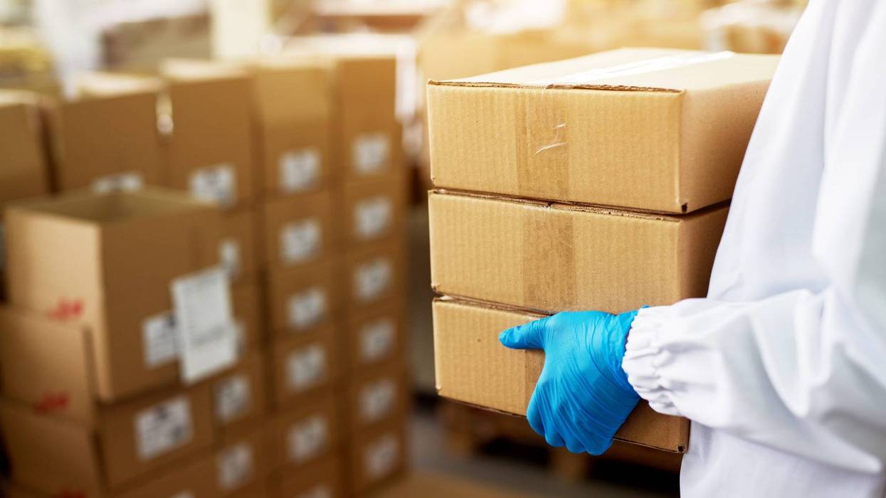 Close up view of a dedicated worker carrying a stack of duck taped brown boxes in factory storage room while wearing sterile cloths and rubber gloves.