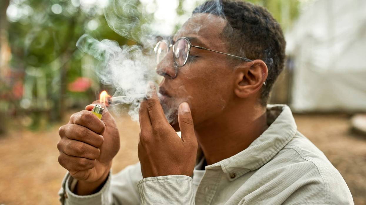 Close view of young afro guy lighting a joint, wearing grey shirt and glasses, smoking on the fresh air , a lot of smoke on the picture, holding a yellow lighter in hand