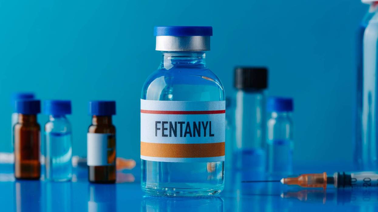 closeup of a simulated vial of fentanyl on a blue table next to a syringe and some other different vials