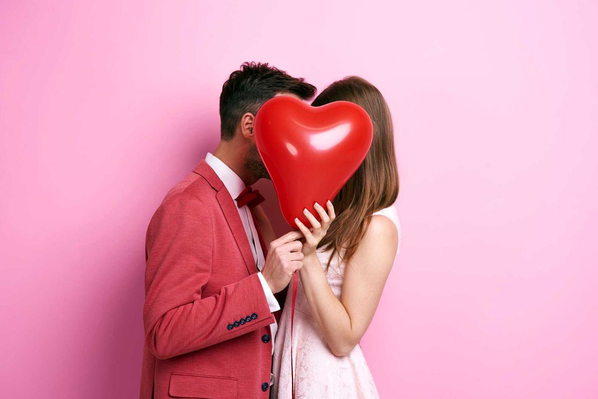 Couple kissing behind a red heart balloon against a pink background.