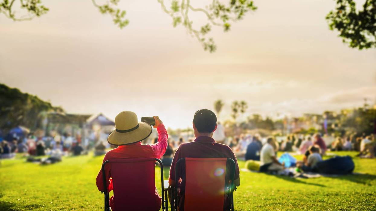 Couple taking photos using smartphone at sunset, unrecognizable crowd of people listen to the music concert in the park.