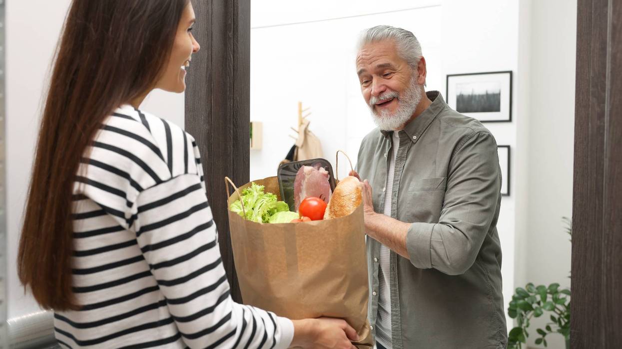 Courier giving paper bag with food products to senior man indoors