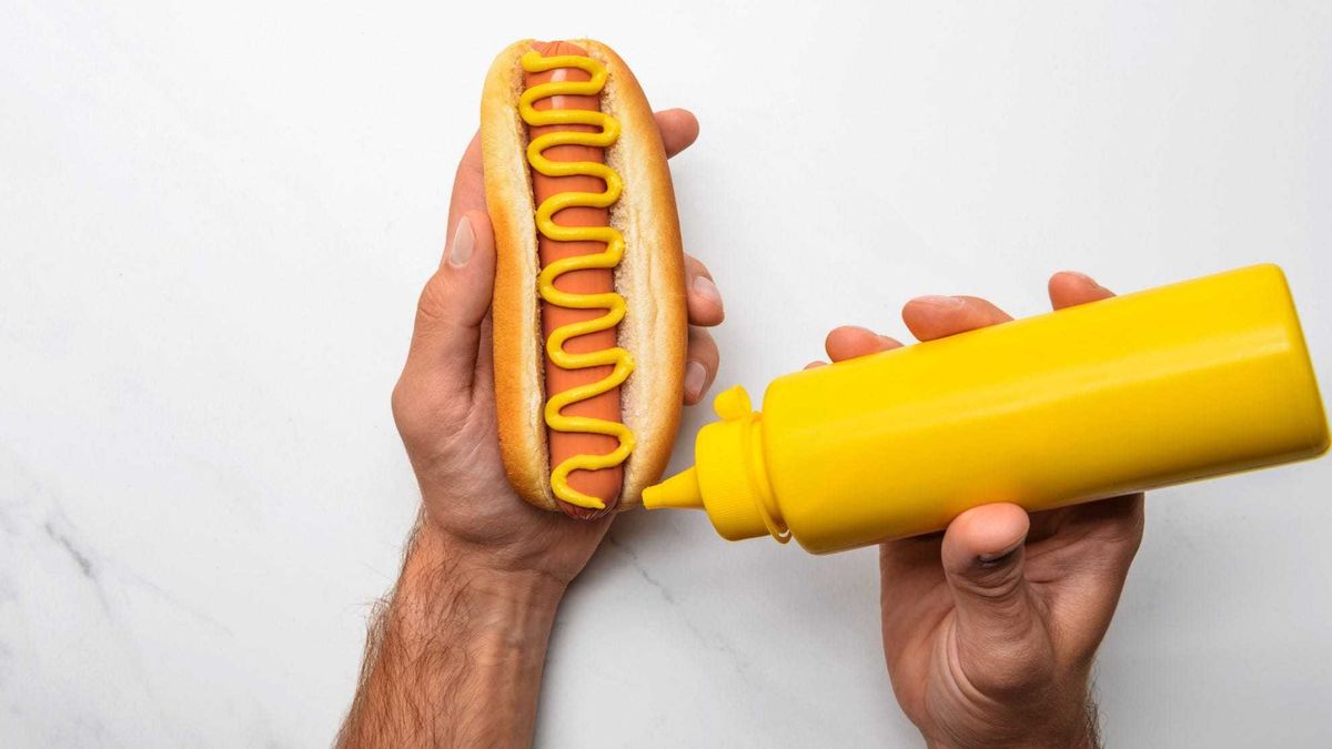 cropped shot of man pouring mustard onto hot dog on white marble surface