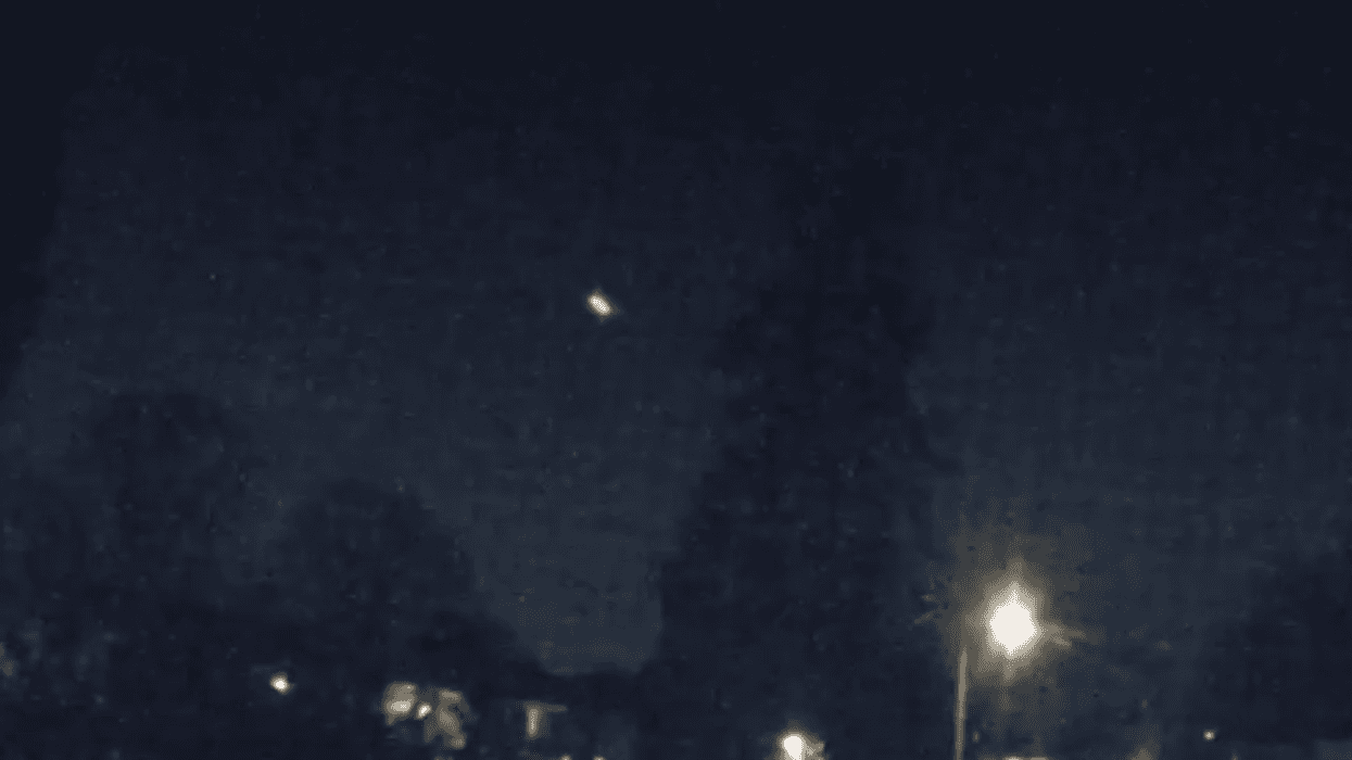 Dark, blurry night scene with a bright, elongated light in the sky above a streetlamp and houses.