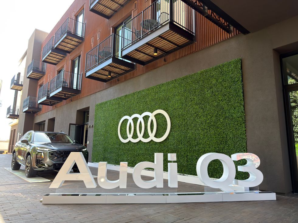 Dark gray Audi Q3 SUV displayed with large 'Audi Q3' text and rings logo on a green hedge wall.