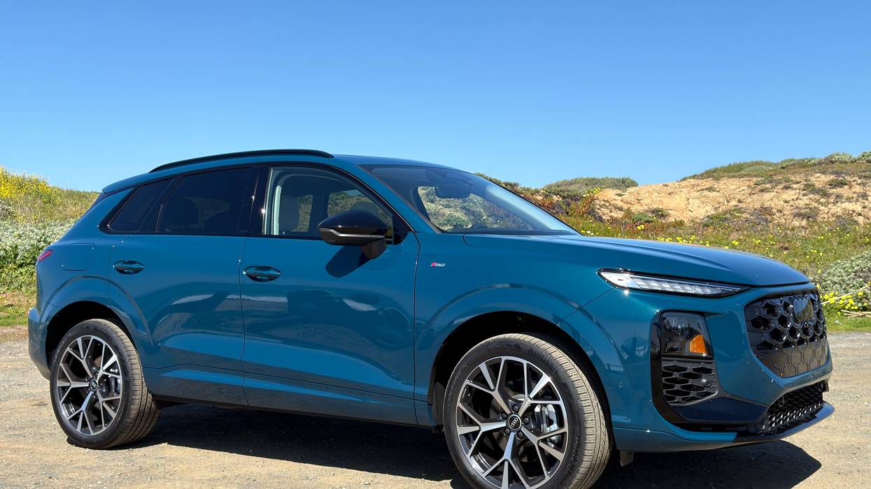 Dark teal Audi SUV on gravel path under sunny blue sky, hills with wildflowers.