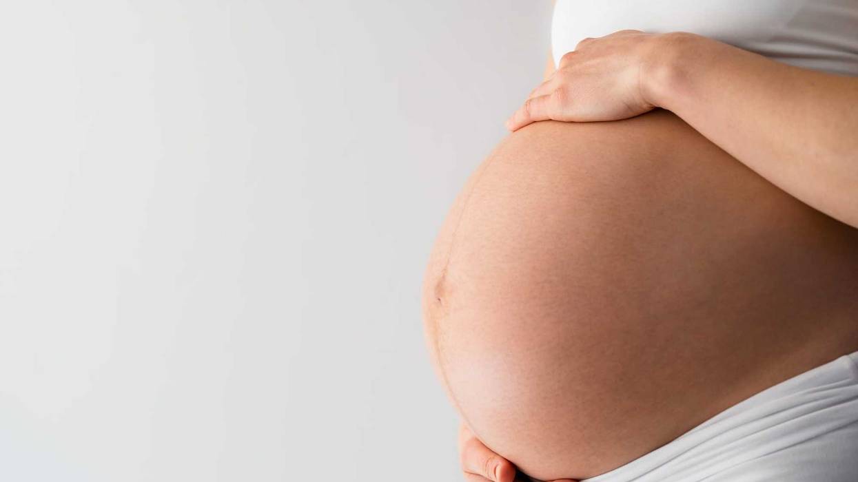 Description: Midsection of unrecognizable standing mother gently holding her very round pregnant baby belly. Side angle view. White background. Bright shot.