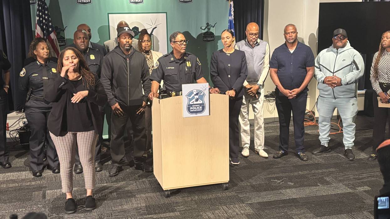 Detroit Police press conference: officer speaking at podium, sign language interpreter, diverse group.