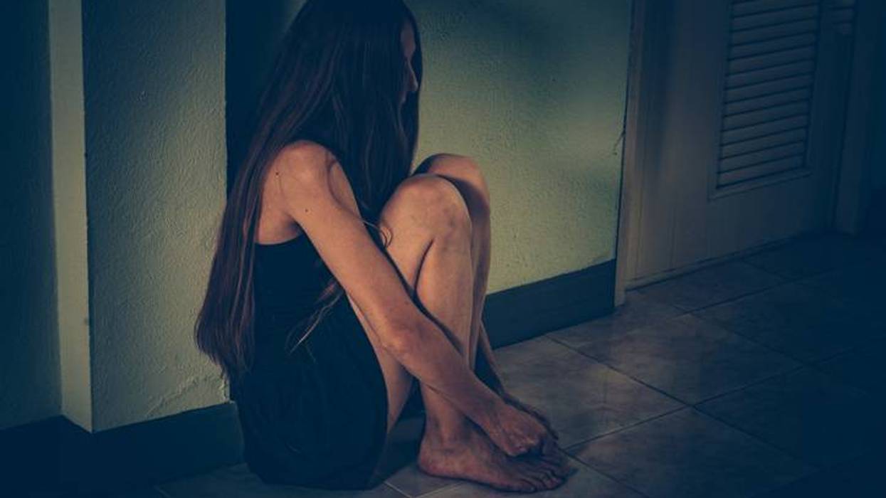 Distressed woman with long hair huddled in a dark corner on tiled floor.