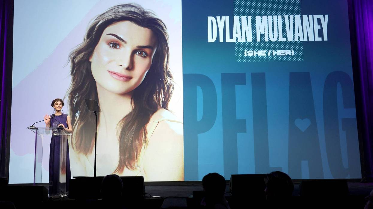 dylan mulvaney speaking onstage w a photo of herself projected on screen behind her