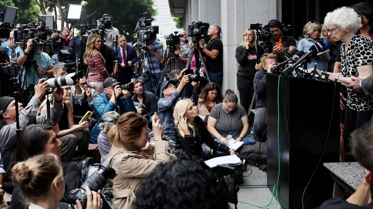 elderly woman speaking in front of crowd of reporters