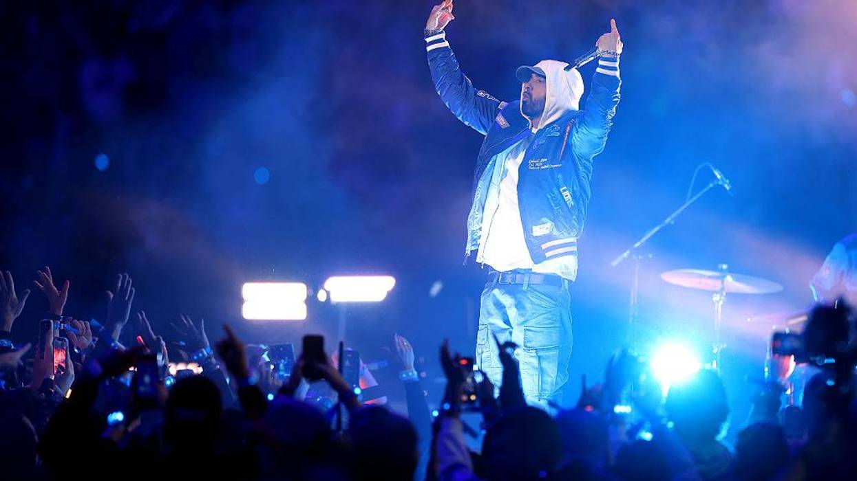 Eminem on stage, arms raised, pointing up, performing live under blue lights to a crowd.