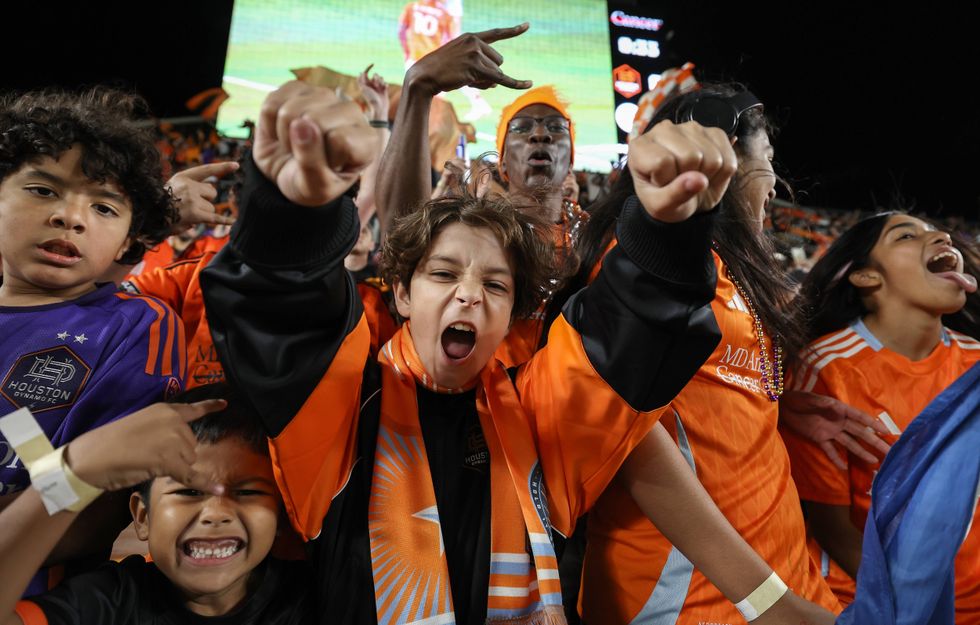 Energetic Houston Dynamo FC fans, diverse kids and adults, cheer wildly in orange at a night game.