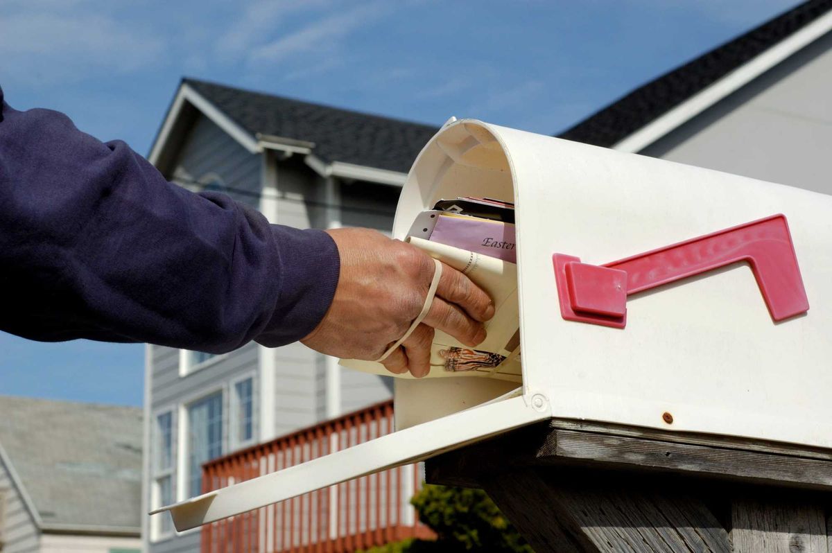 File - Mailperson's arm inserting a bundle of mail into a mailbox. Partially obscured suburban home in background.