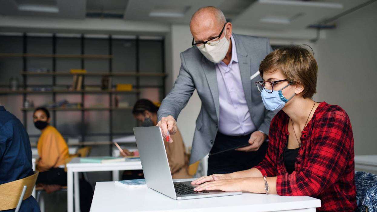 File photo of a masked teacher helping a masked student at her desk.