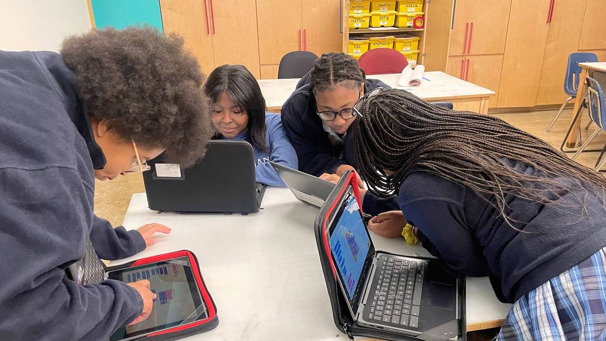 four girls sit at a table with laptops and tablets. they are all black and wear their school uniforms which are navy blue, and are intently focused on their work.