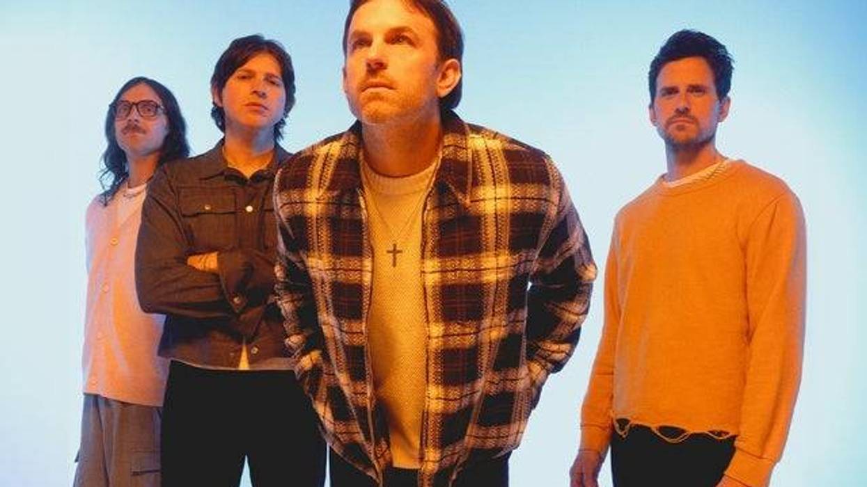 Four men in casual clothes stand against a light blue background, looking serious.
