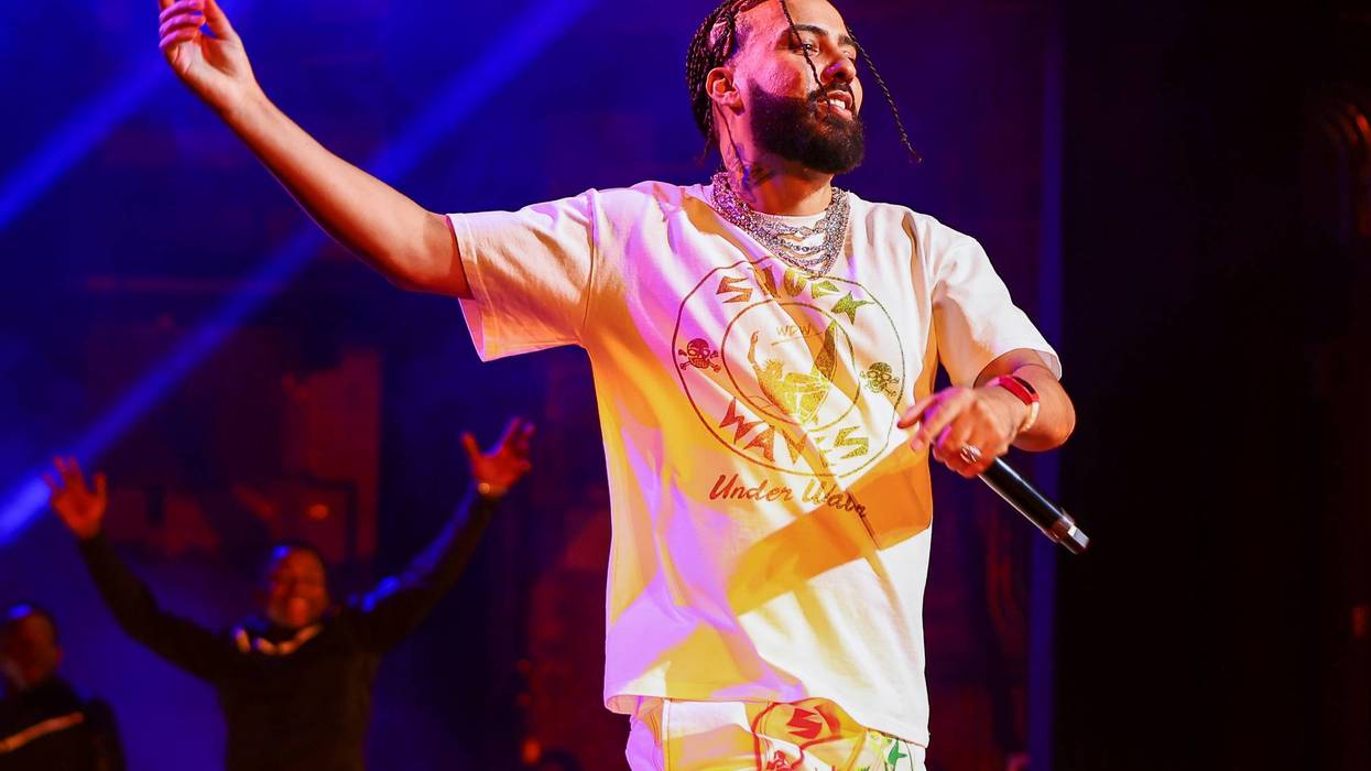 French Montana performs on stage, arm raised, holding a microphone, under blue and yellow lights.