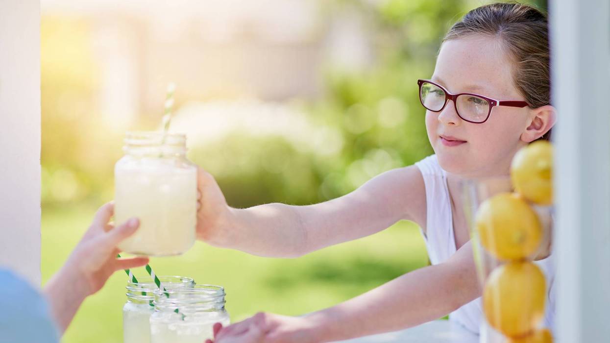 Girl in glasses hands a jar of lemonade to a customer at an outdoor stand with lemons.