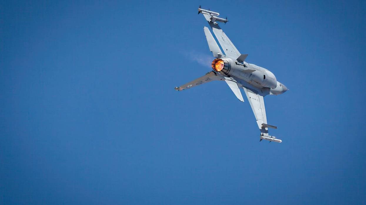 Gray fighter jet banking sharply, afterburner flaming orange against a clear blue sky.