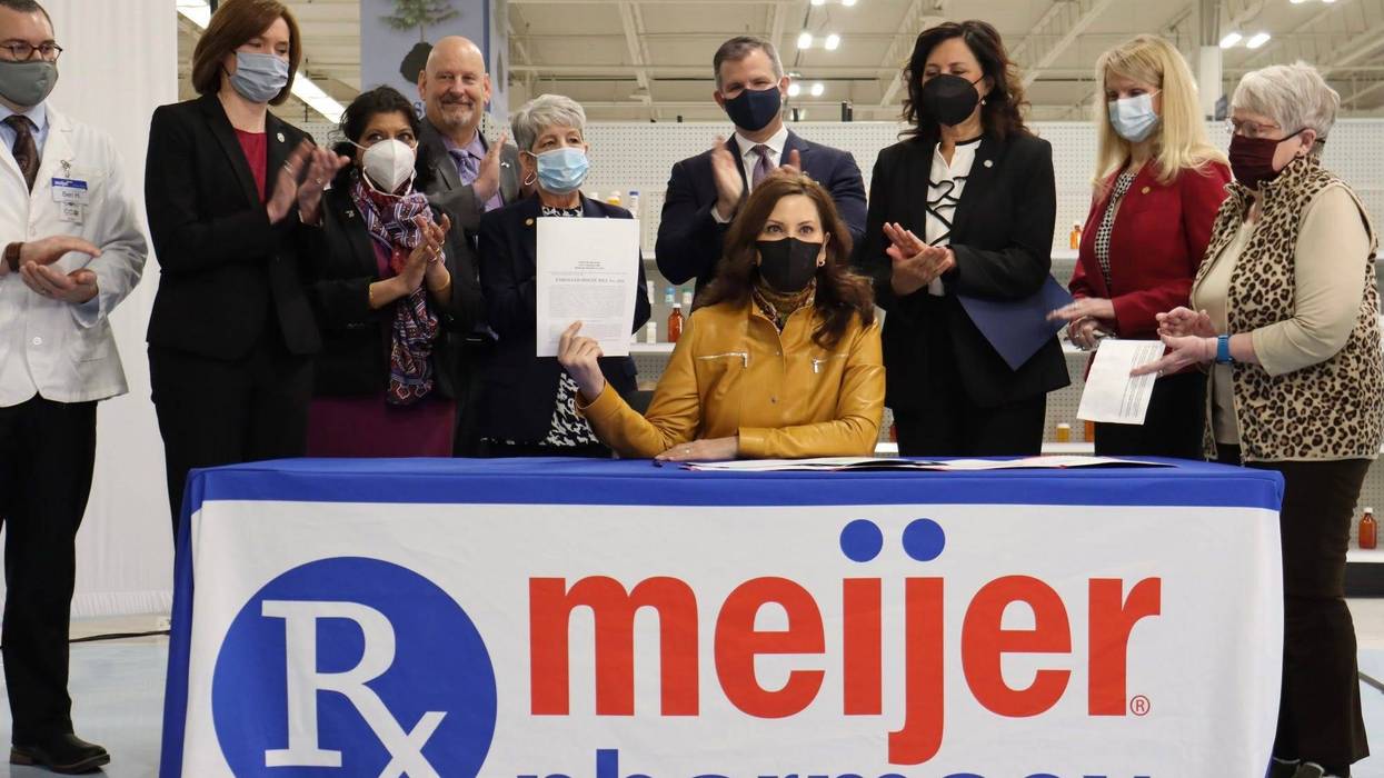Gretchen Whitmer holding up signed bill sitting at table that says Meijer pharmacy and surrounded by people standing and clapping