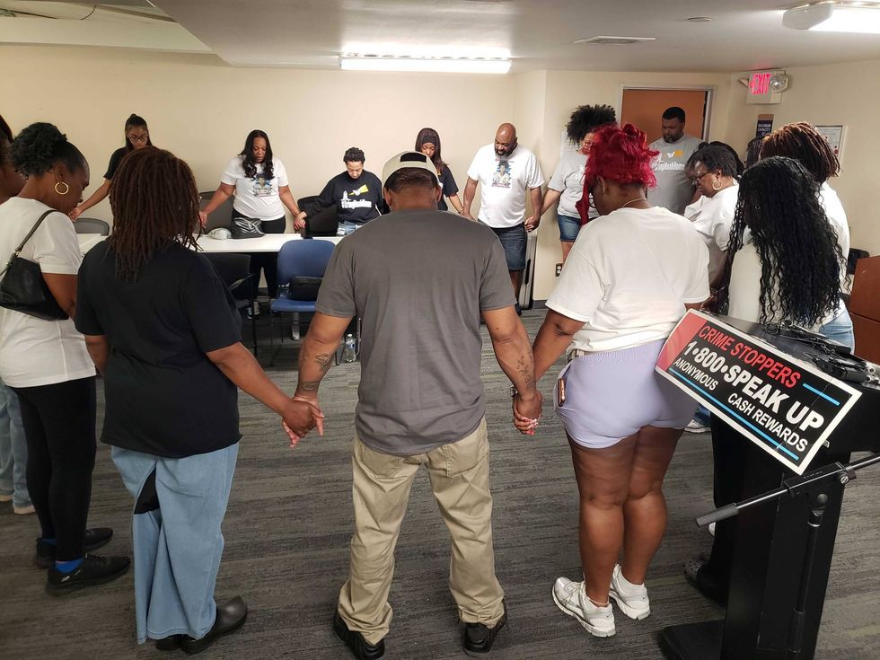 Group of adults holding hands, heads bowed in solidarity or prayer, near a Crime Stoppers sign.