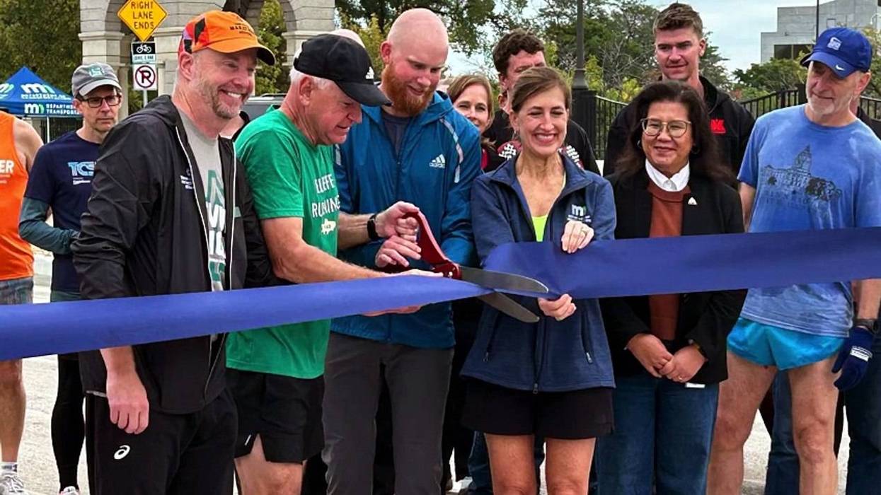 Group of smiling runners and officials cutting a blue ribbon outdoors.