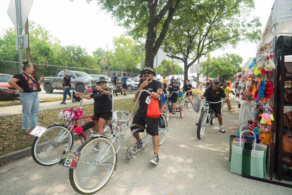 Group riding customized lowrider bikes at a vibrant street festival with colorful vendor stalls.