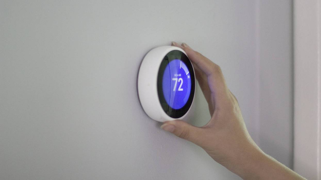 Hand adjusting a white smart thermostat displaying 72 on a grey wall.
