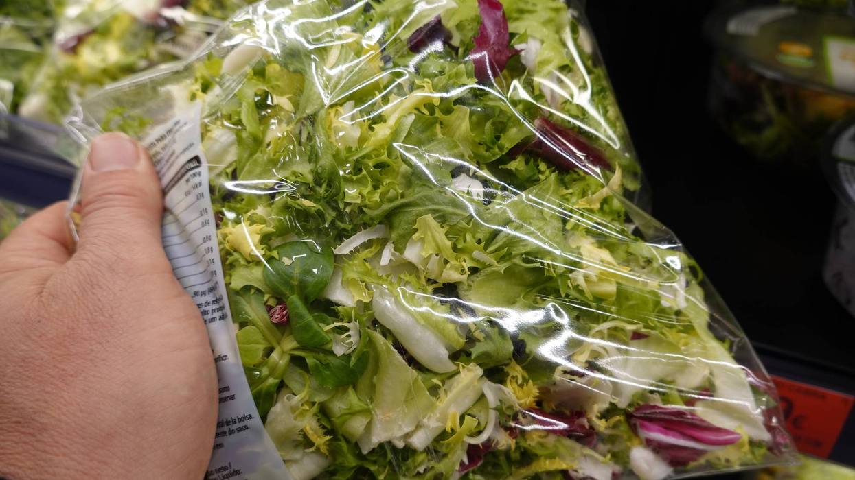 Hand holding a clear plastic bag of mixed green and red lettuce in a supermarket.