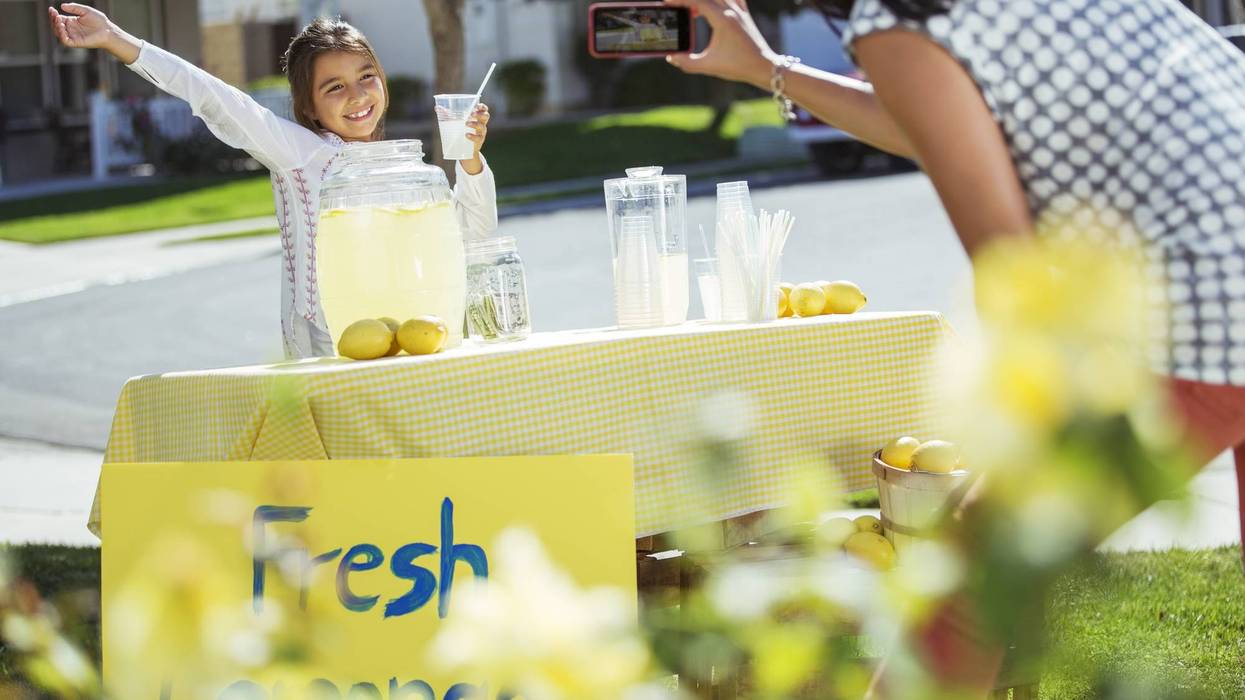 Happy girl poses for a photo at her lemonade stand with a 'Fresh Lemonade' sign.