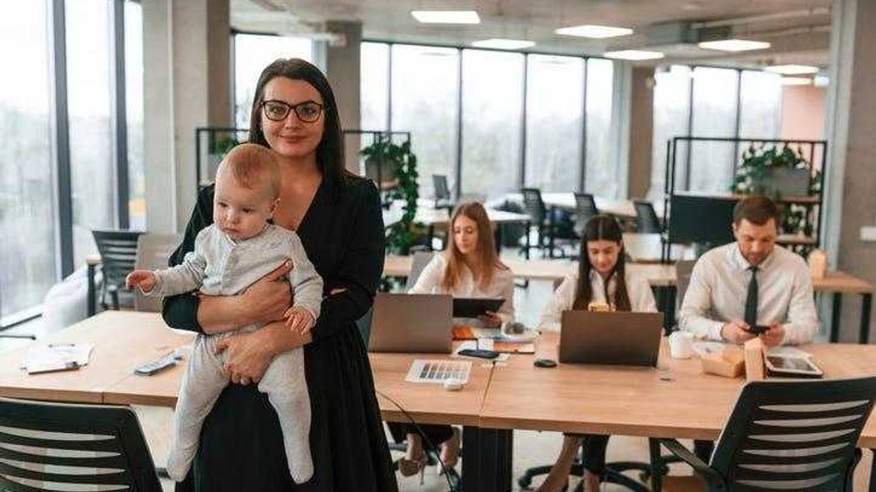 Happy mother is standing and holding her little son. Infant baby is in the office where group of people are working together.