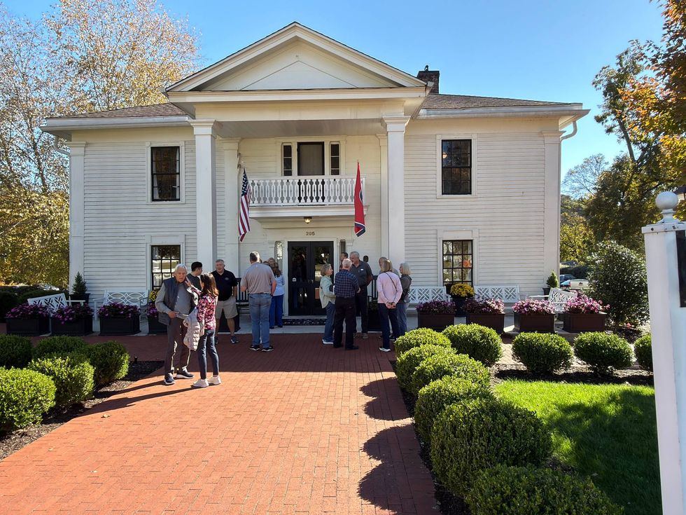 Historic white building with columns and two flags, surrounded by lush greenery and flowers. A group of people is gathered outside on a sunny day.