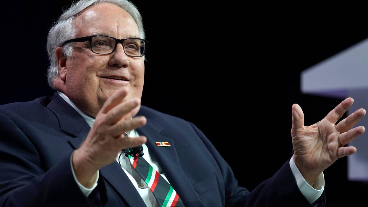 howard buffett photo. he is speking on a stage sitting in a chair gesturing with his hands and smiling. he has gray hair and glasses and a navy blue suit on
