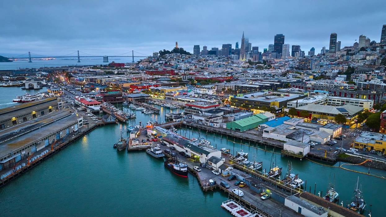 Image of Fishermans Wharf at dusk with night lights on over city and pier with Oakland Bay Bridge aerial