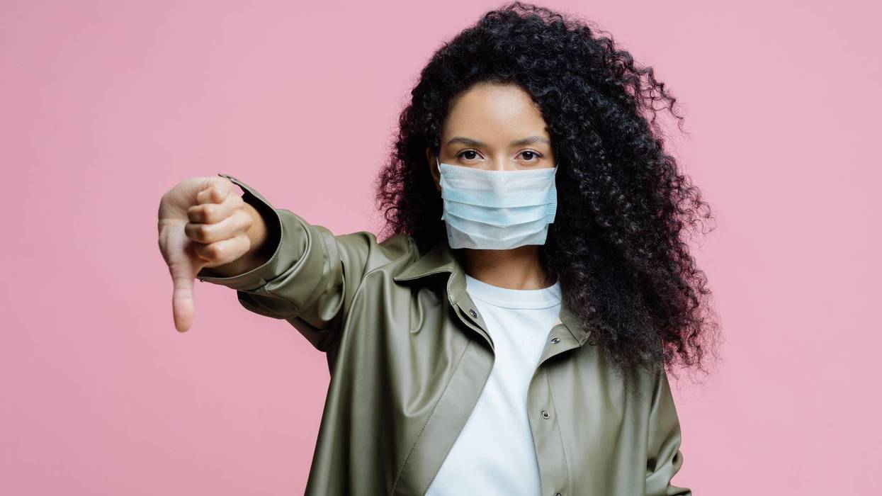 Image of serious curly haired young woman wears medical mask, keeps thumb down, shows dislike gesture, avoids spreading of corona virus, cares about health.