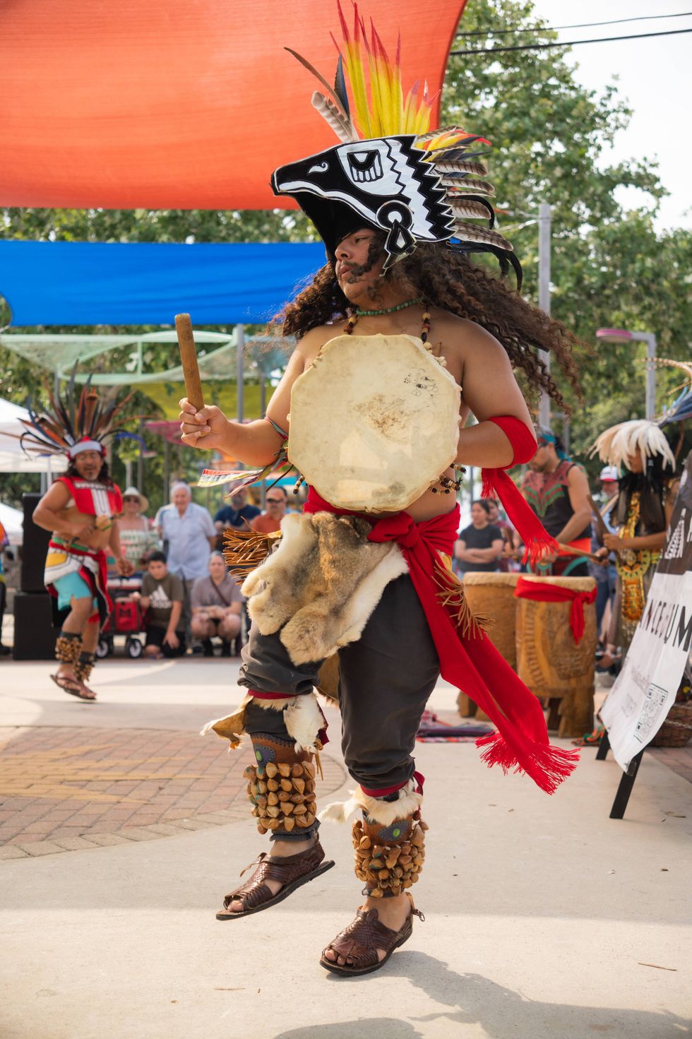 Indigenous dancer in ornate feathered headdress, traditional clothing, drumming outside.