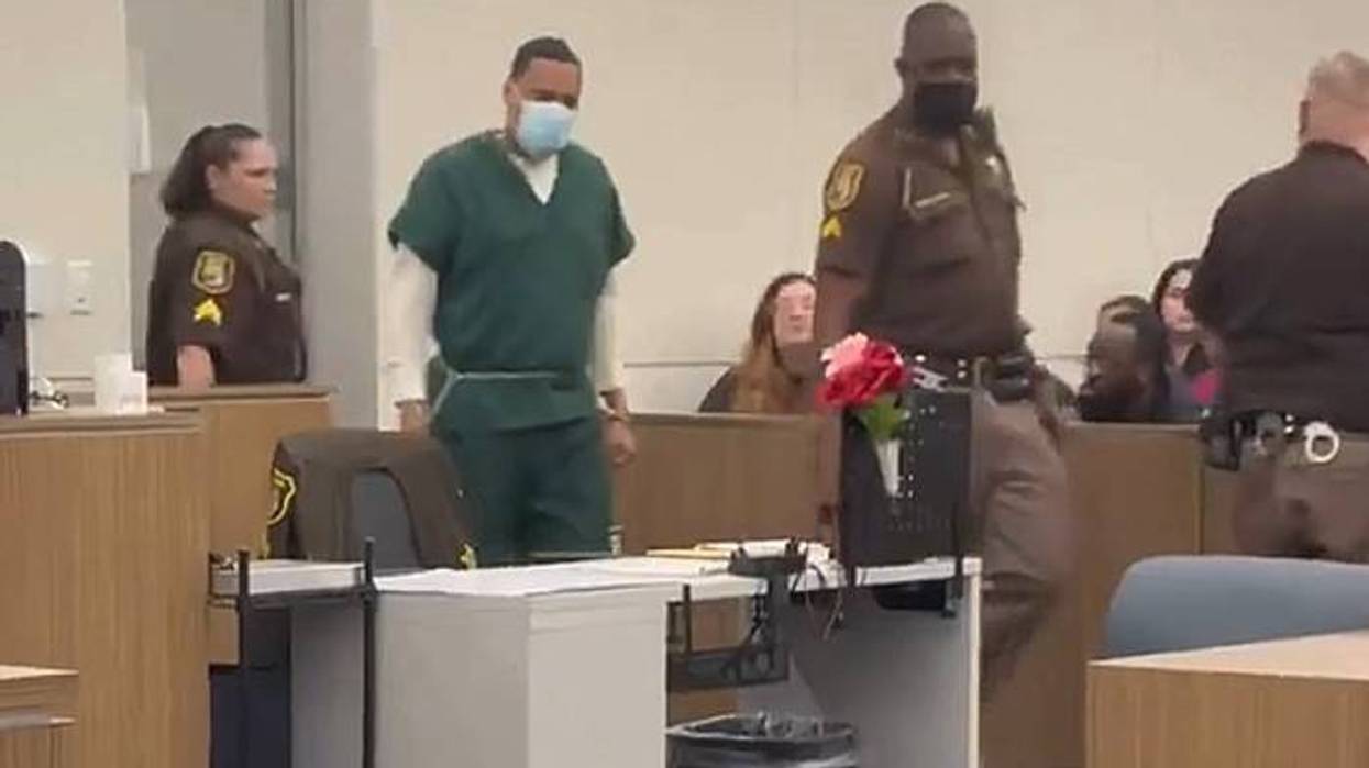 Inmate in green scrubs and mask escorted by deputies in courtroom; flowers on desk.