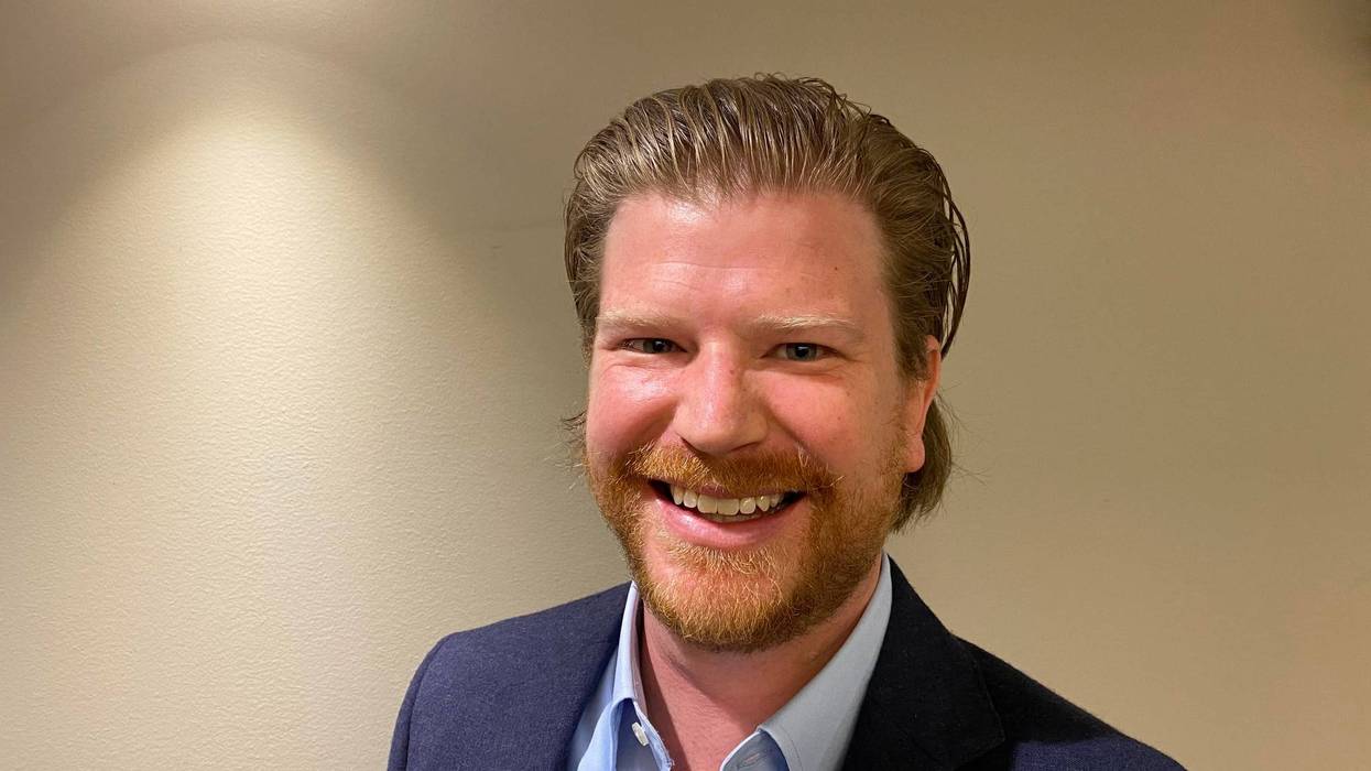 Jack Coatar stands smiling. he has dirty blond hair and beard and blue suit on