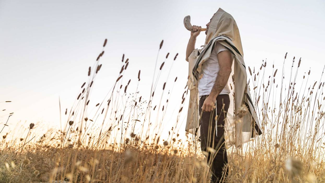 Jewish man in a traditional Tallit prayer shawl blowing the ram's horn Shofar, in the field against sunrise sky on Rosh Hashana and Yom Kippur.