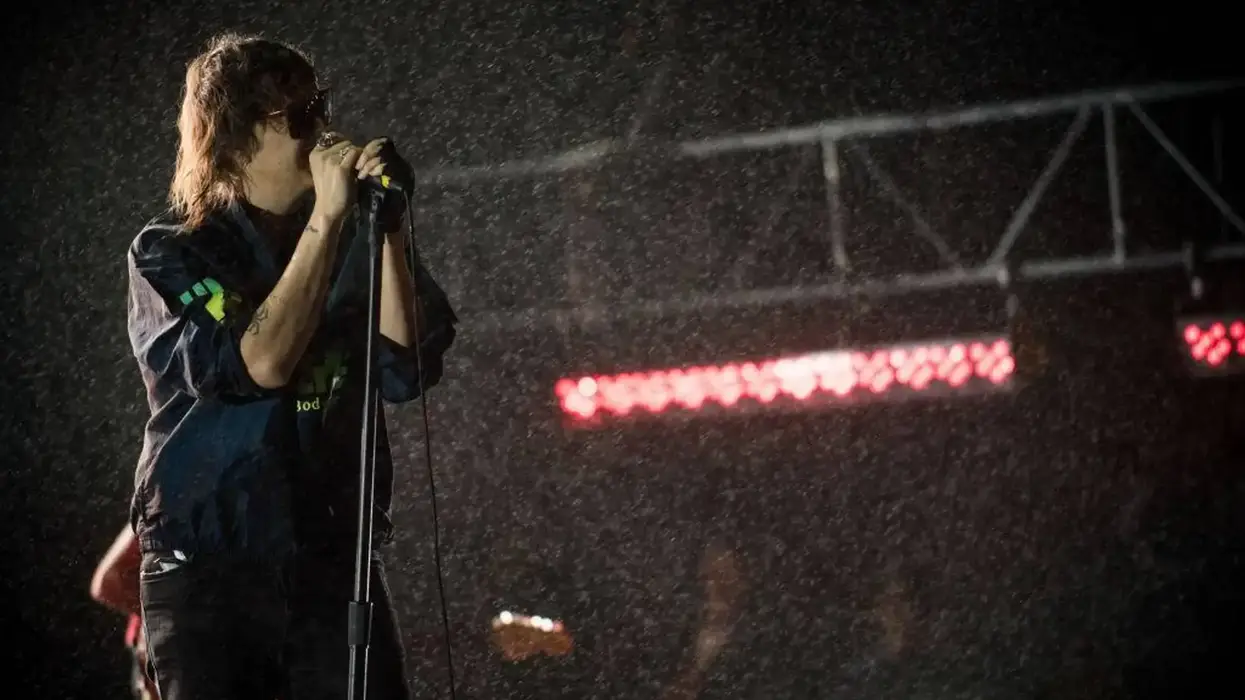 Julian Casablancas sings into a microphone, rain-soaked on a dark stage with red lights.