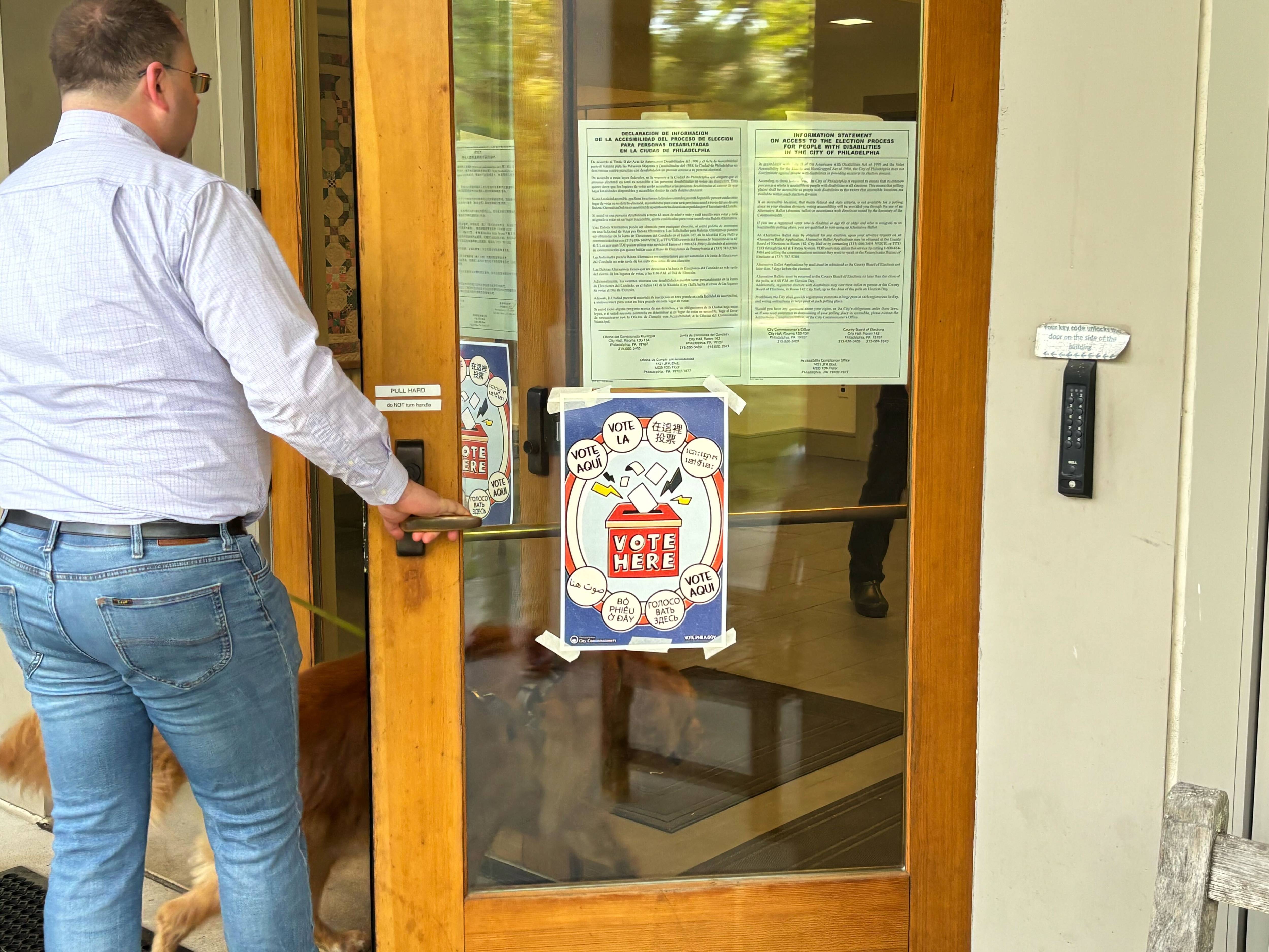 Man and dog pull open a glass door marked 'Vote Here,' entering a polling place.